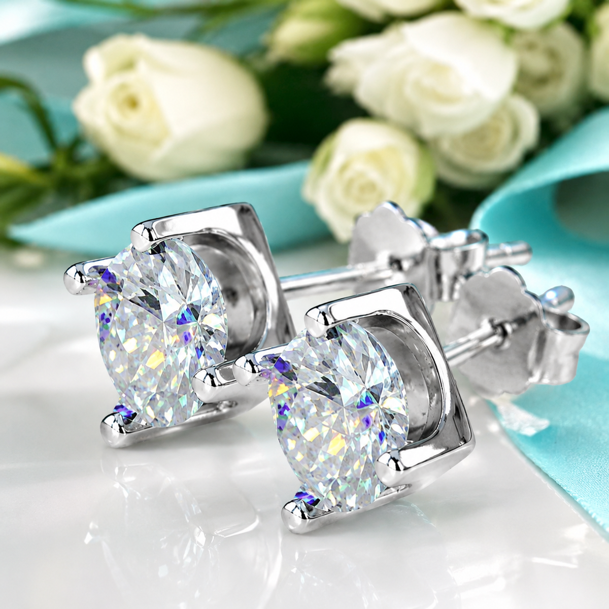 A pair of silver stud earrings with large oval clear gemstones, displayed on a glossy white surface with white roses and a light blue ribbon in the background.