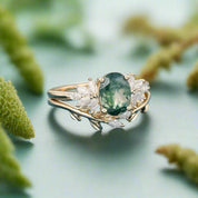 Gold ring with a green gemstone and diamonds on a blue surface with blurred dried flowers in the background