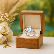 Engagement ring with emerald-cut gemstone in a wooden box, surrounded by flowers.