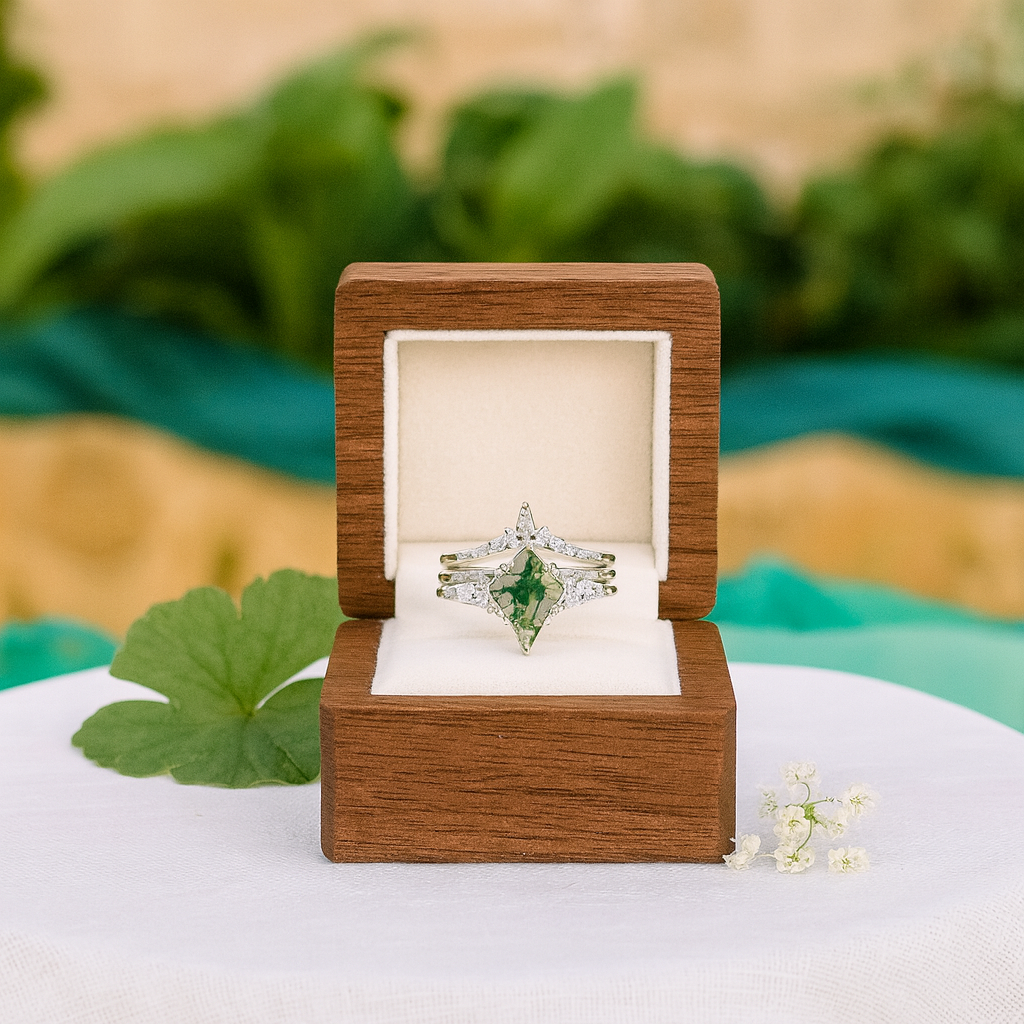 Engagement ring with green gemstone in a wooden box on a white surface with green leaves.