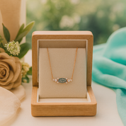 Gold necklace with a green stone in a wooden jewelry box, surrounded by flowers and fabric.