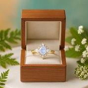 Gold ring with a large central gemstone in a wooden box, surrounded by greenery and flowers.