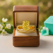 Gold ring with a large yellow gemstone in a wooden box, surrounded by flowers and a green object.