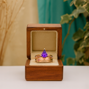 Gold ring with a purple gemstone in a wooden box on a white surface with a blurred green plant background.
