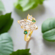 Gold ring with large clear gemstone and smaller green gemstones on a beige background