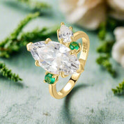 Gold ring with large clear gemstone and smaller green gemstones on a white background