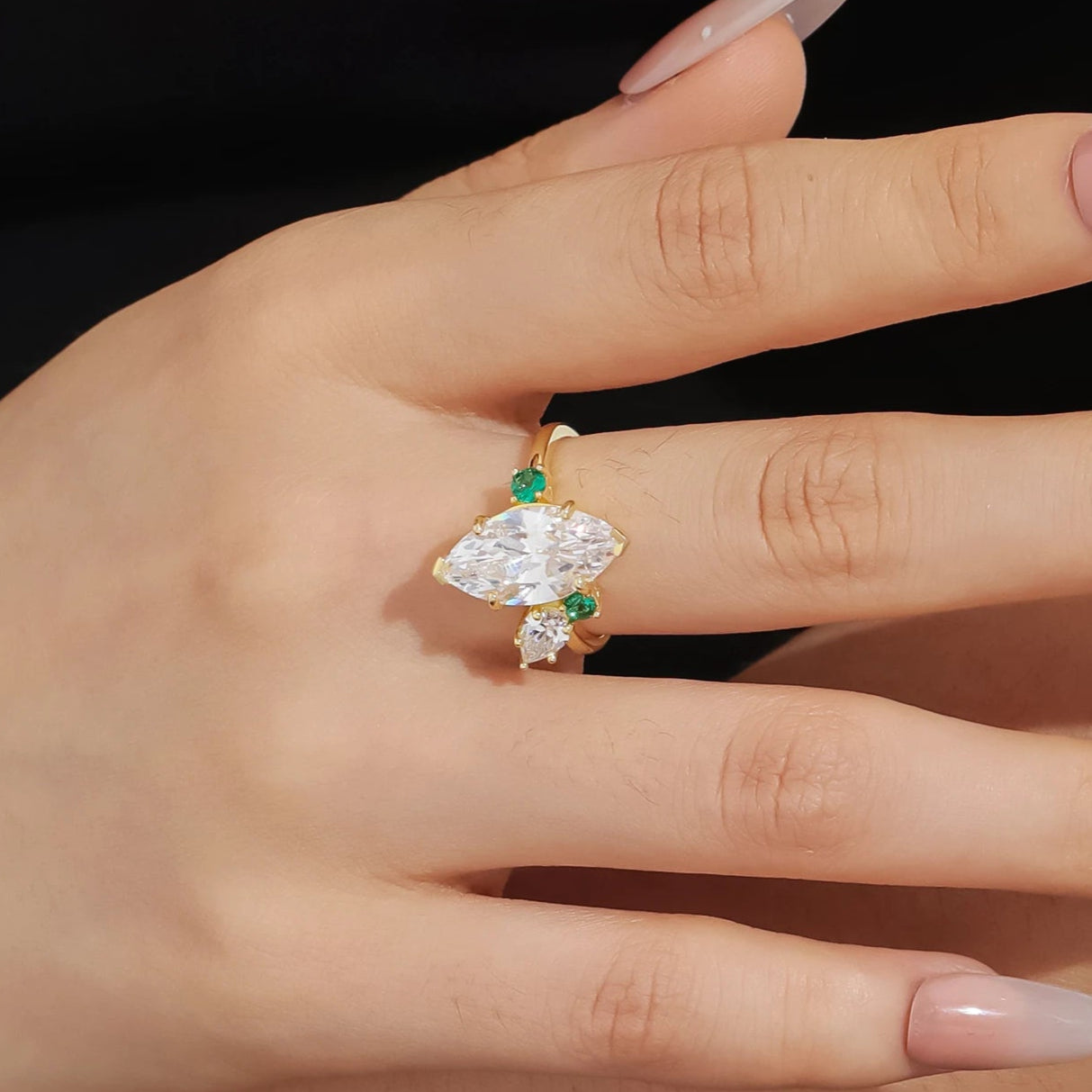 Hand wearing a gold ring with emerald and diamond stones against a black background
