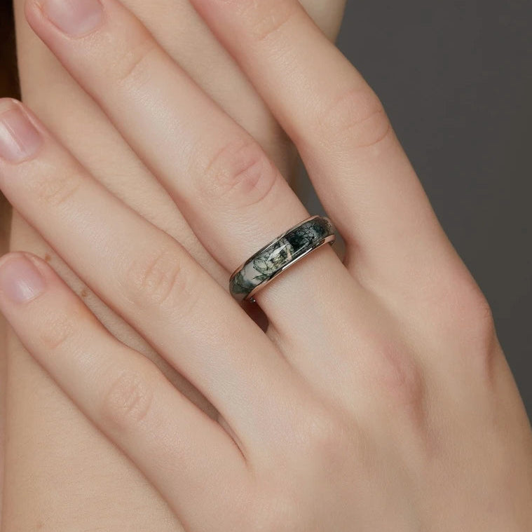 Hand wearing a ring with a dark band design, against a neutral background.