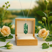 Necklace with a green pendant in an open wooden box, surrounded by flowers outdoors.
