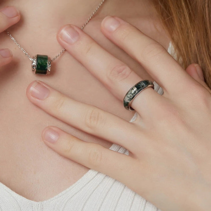 Person wearing a silver necklace with a green gemstone and a matching ring on a plain background