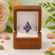 Ring with a purple gemstone in a wooden box on a light surface with flowers and leaves in the background