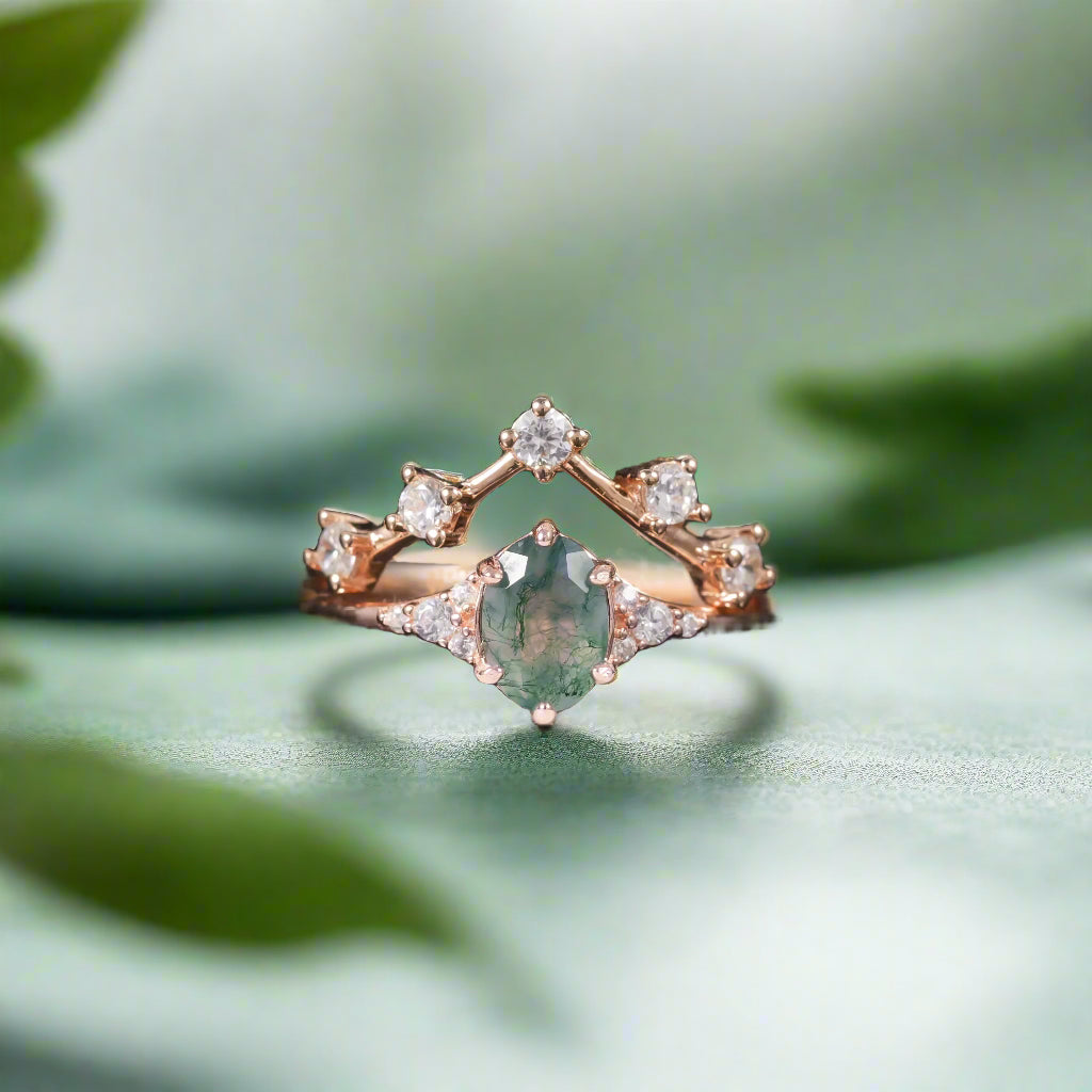 Rose gold ring with a green gemstone and diamonds on a dark surface with blurred green leaves in the background