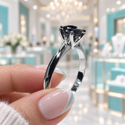 A hand with French-tipped nails holds a silver-toned solitaire ring featuring a black gemstone in a bright, blurred jewelry store setting with turquoise accents.