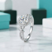 A silver engagement ring with a round-cut center stone and a halo of smaller stones, displayed on a light marble surface with blurred turquoise and white gift boxes in the background.