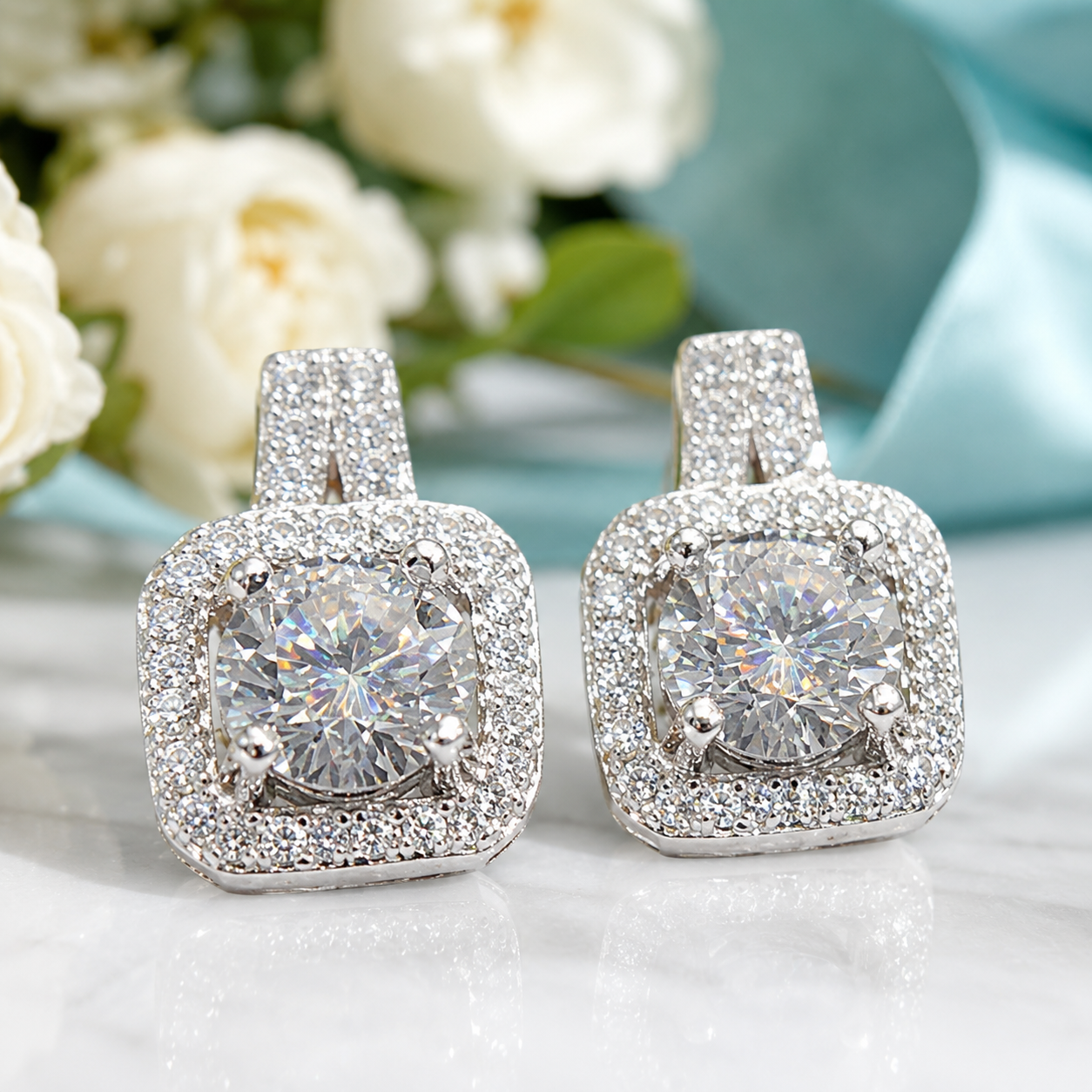 A pair of square halo stud earrings with large round clear center stones and surrounding pavé settings, displayed on a light reflective surface with soft white flowers and a pale blue fabric in the background.
