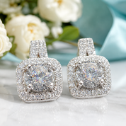 A pair of square halo stud earrings with large round clear center stones and surrounding pavé settings, displayed on a light reflective surface with soft white flowers and a pale blue fabric in the background.