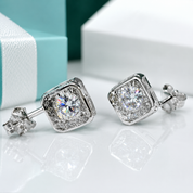 A pair of silver-toned square stud earrings with clear gemstones, displayed on a reflective white surface with a light blue and white gift box in the background.