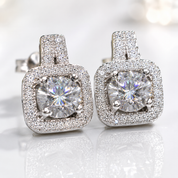 A pair of square cushion-cut halo stud earrings featuring brilliant round center stones surrounded by a double row of pavé-set clear stones in a silver-toned setting, displayed on a reflective white surface.