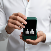 A person in a white button-up shirt holds an open dark green jewelry box displaying a pair of square-shaped silver-toned earrings with clear stones.