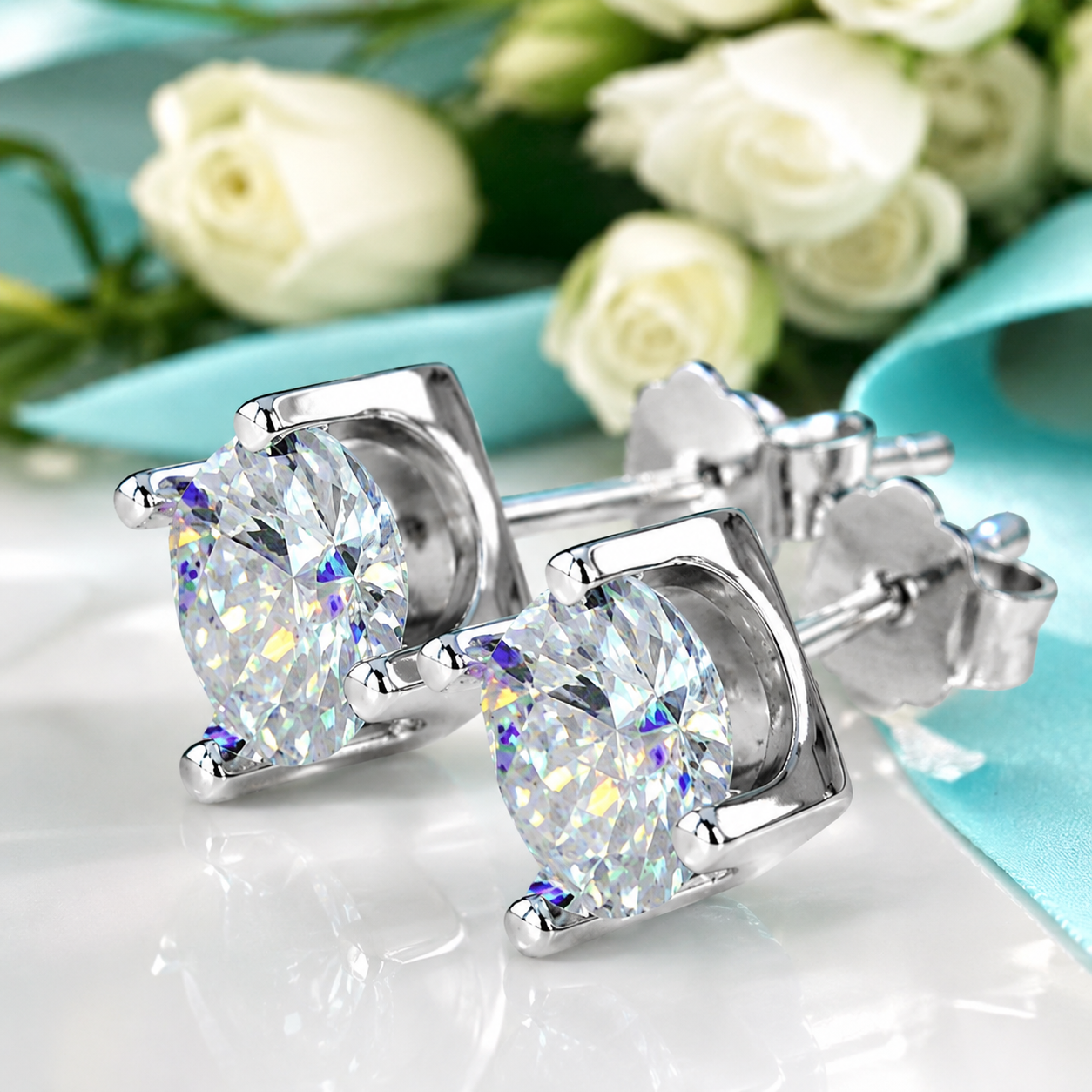 A pair of silver stud earrings with large oval clear gemstones, displayed on a glossy white surface with white roses and a light blue ribbon in the background.