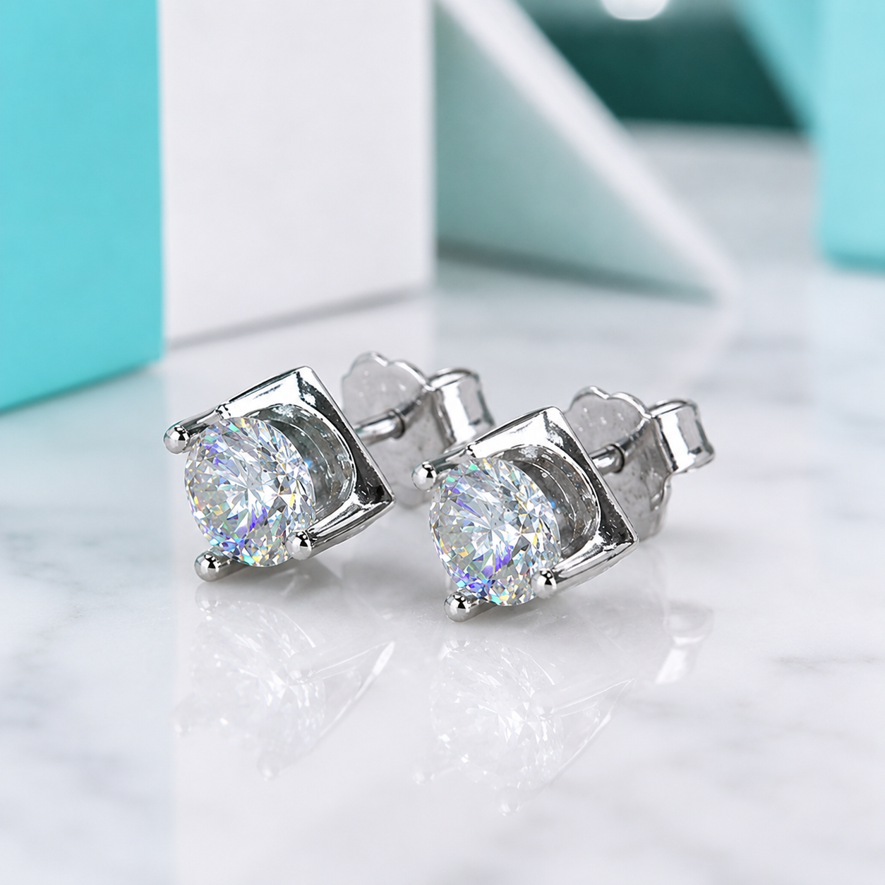 A pair of silver stud earrings with large round clear gemstones, displayed on a white marble surface with a blurred turquoise and white background.