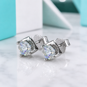 A pair of silver stud earrings with large round clear gemstones, displayed on a white marble surface with a blurred turquoise and white background.