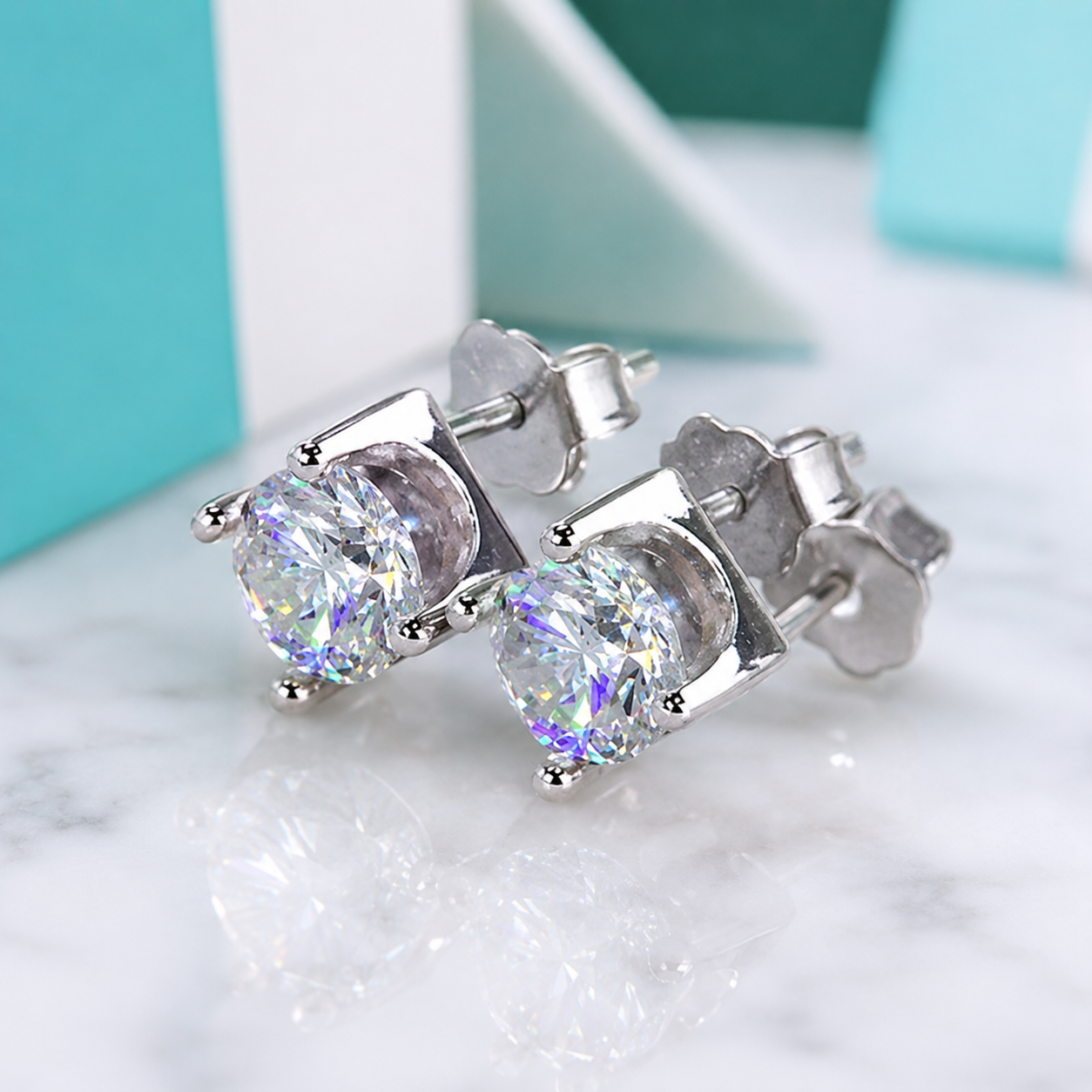 A pair of silver stud earrings with large round iridescent clear gemstones, displayed on a white marble surface with a blurred turquoise and white background.