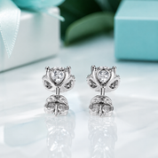 A pair of silver-toned stud earrings with heart-shaped clear gemstones, displayed on a glossy white surface with soft-focus gift boxes and green leaves in the background.