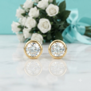 A pair of round bezel-set stud earrings with clear gemstones in yellow gold settings, displayed on a white marble surface with a reflection. In the background, a bouquet of white roses with a light blue ribbon is softly blurred against a light blue backdrop.