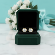 A pair of round gold stud earrings with clear gemstones displayed in an open dark green jewelry box on a white marble surface, with blurred white flowers and a light blue ribbon in the background.