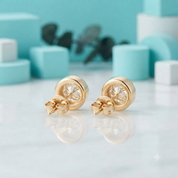 A pair of gold stud earrings with clear round-cut gemstones, displayed on a reflective white surface with a soft-focus turquoise and white geometric background.