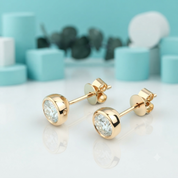 A pair of gold stud earrings with round bezel-set clear gemstones, displayed on a reflective white surface with a soft-focus turquoise and white background.