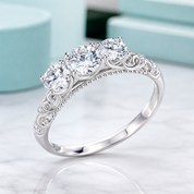 A silver three-stone diamond ring with ornate scrollwork on the band, displayed on a light marble surface with a soft-focus turquoise background.