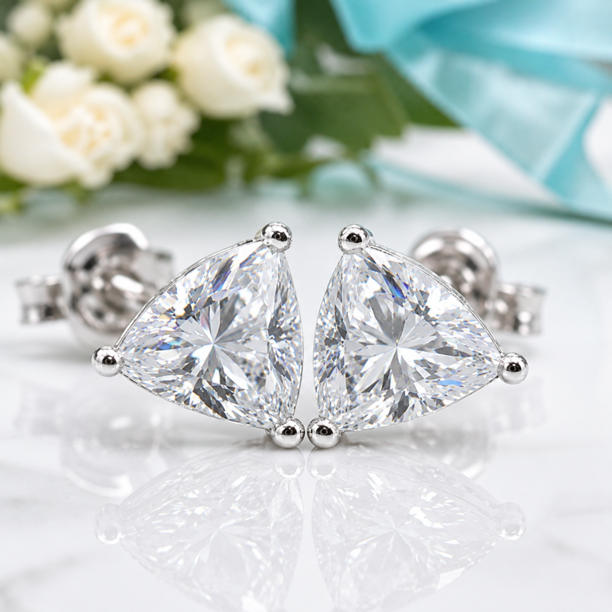 A pair of triangular diamond stud earrings in silver settings, displayed on a reflective white surface with soft-focus white roses and a light blue ribbon in the background.