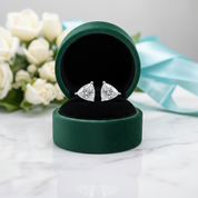 A pair of triangular diamond stud earrings displayed in an open dark green velvet jewelry box on a white marble surface, with white roses and a light blue ribbon blurred in the background.