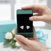 A pair of triangular stud earrings with clear gemstones set in silver-toned metal, displayed inside an open dark green jewelry box held by two hands. A light blue jewelry box and ribbon are blurred in the background, along with white flowers.
