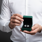 A person in a white dress shirt holds an open dark green jewelry box displaying a pair of silver-toned cufflinks on a black velvet insert against a dark gray background.