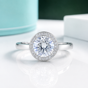 A round halo engagement ring with a brilliant-cut center stone and a halo of smaller stones, set in a polished silver-colored band. The ring is displayed on a clean white surface with a soft reflection, featuring a blurred turquoise and white background.