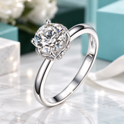 A silver solitaire engagement ring with a round brilliant-cut diamond and a halo of small accent diamonds, displayed on a white marble surface with soft-focus gift boxes in the background.