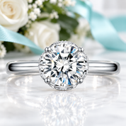 A classic solitaire engagement ring featuring a round brilliant-cut diamond set in a silver-toned four-prong setting, resting on a white marble surface with a soft reflection. The background is a soft-focus floral arrangement with white roses and a light blue ribbon.