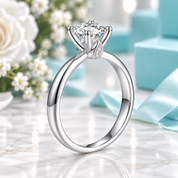 A silver-toned solitaire engagement ring with a large round brilliant-cut diamond and a small accent stone, displayed on a marble surface with light blue gift boxes and white flowers in the background.