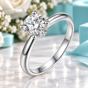 A silver solitaire engagement ring with a round brilliant-cut diamond set in a four-prong setting, resting on a reflective marble surface with soft-focus turquoise gift boxes and white flowers in the background.