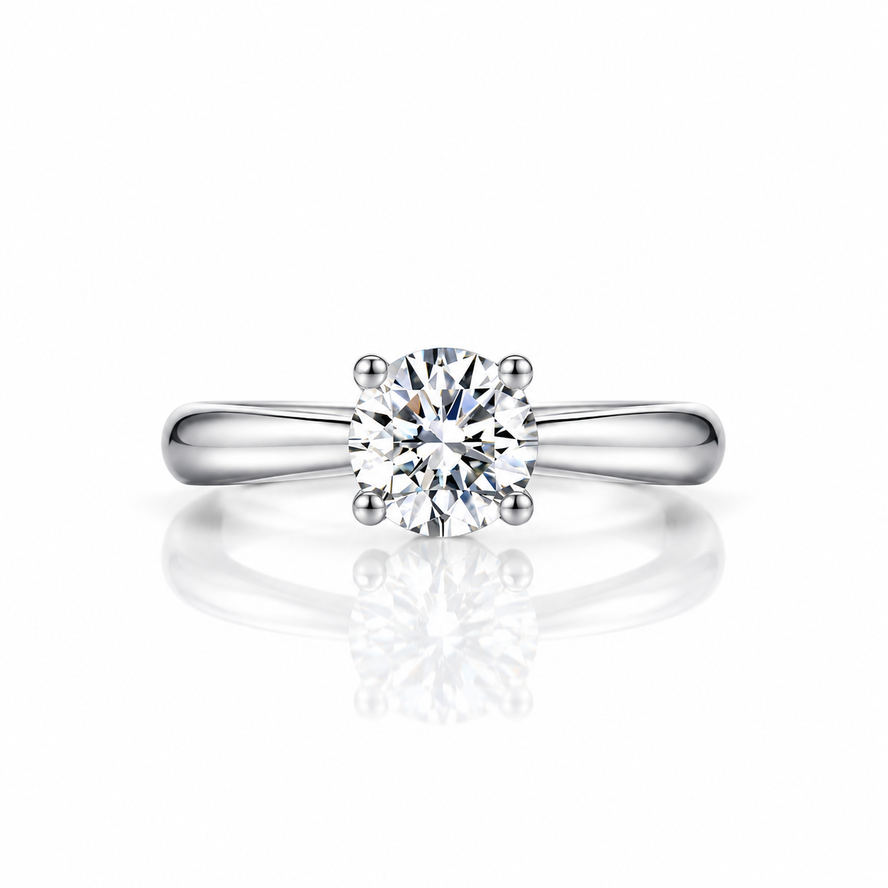A classic solitaire engagement ring featuring a round brilliant-cut diamond set in a polished four-prong white metal band, shown on a clean white background with a soft reflection below.