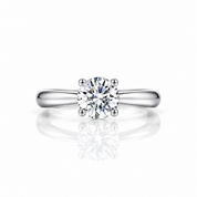 A classic solitaire engagement ring featuring a round brilliant-cut diamond set in a polished four-prong white metal band, shown on a clean white background with a soft reflection below.