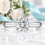 A classic solitaire engagement ring with a round brilliant-cut diamond set in a polished silver-colored band, resting on a white marble surface with a soft reflection. The background is a soft-focus floral arrangement with white roses and a light blue ribbon.