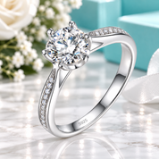 A silver solitaire engagement ring with a round-cut clear gemstone and pavé-set accent stones on the band, displayed on a marble surface with soft floral and turquoise background elements.