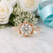 A rose gold engagement ring with a large round-cut clear center stone and a halo setting, accented with small pavé stones on the band, displayed on a light marble surface with white roses, greenery, and a light blue ribbon in the background.