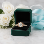 A rose gold engagement ring with a large round-cut center stone and pavé side stones, displayed in an open dark green velvet ring box on a white marble surface, with white roses and a light blue ribbon in the background.