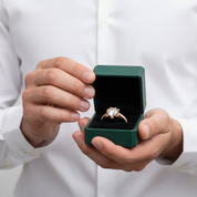 A close-up of a person in a white dress shirt holding an open dark green jewelry box with a gold ring featuring a clear center stone inside.
