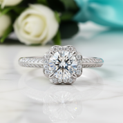 A silver-toned halo ring with a round brilliant-cut center stone and smaller surrounding stones, displayed on a light marble surface with blurred white roses and a teal ribbon in the background.